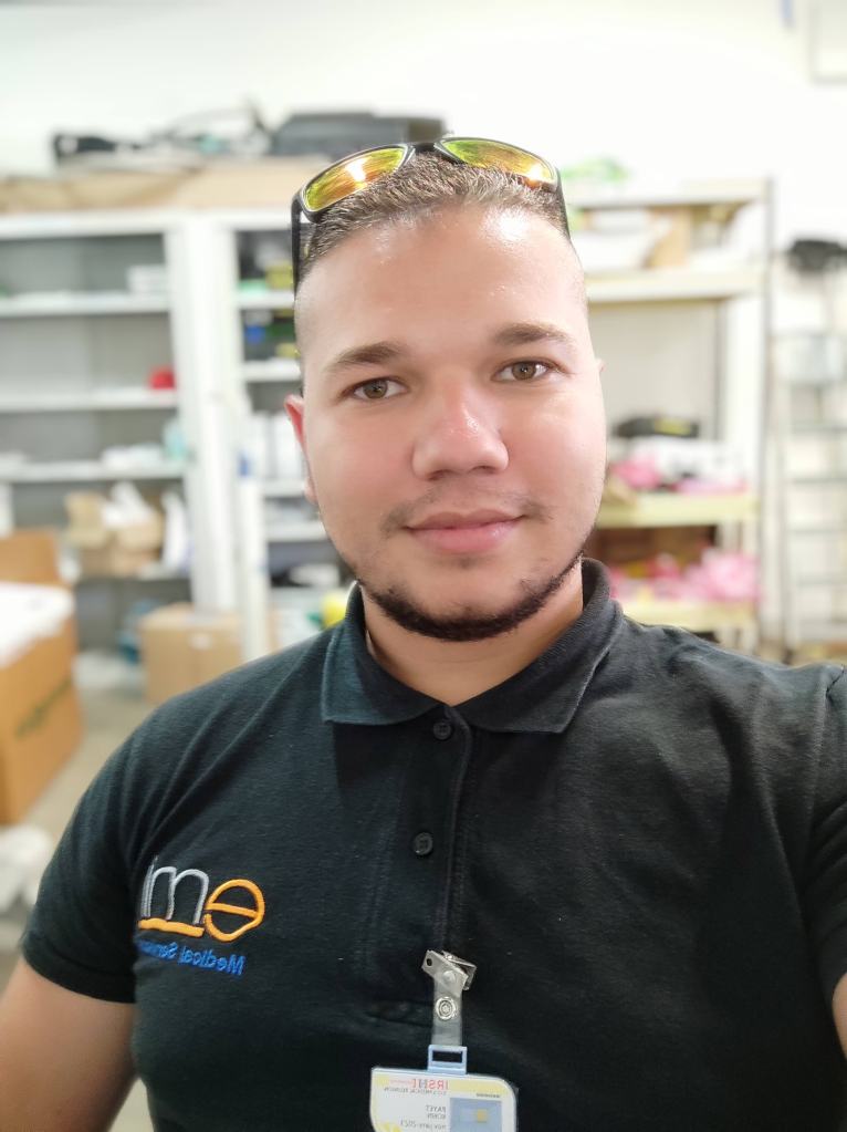 Portrait of a man with sunglasses on his head, wearing a black polo shirt with the logo 'eme'. He is in a workspace with shelves behind him.